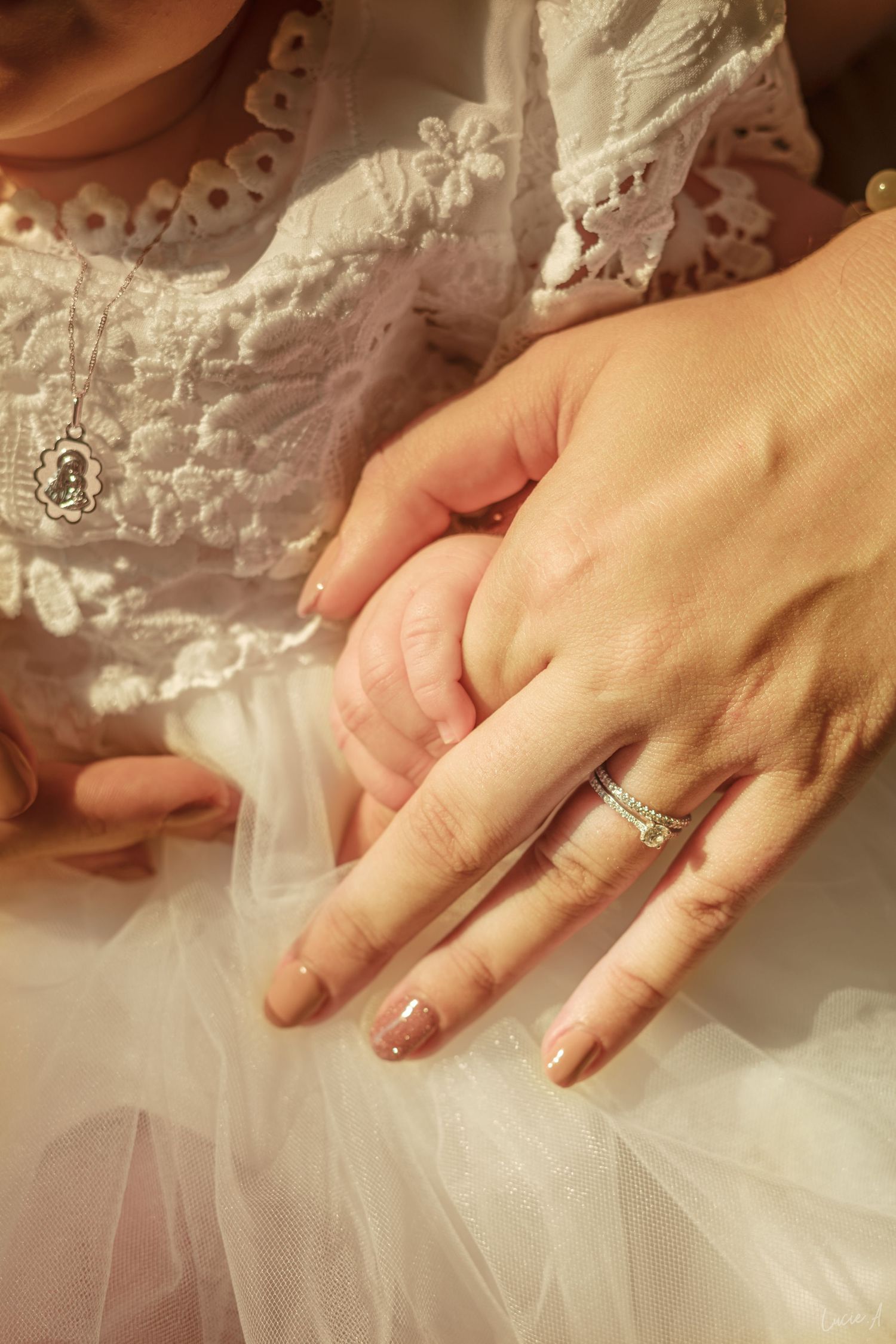Adult hand with diamond ring gently holds baby's hand, both dressed in white lace, symbolizing a tender mother-child bond.