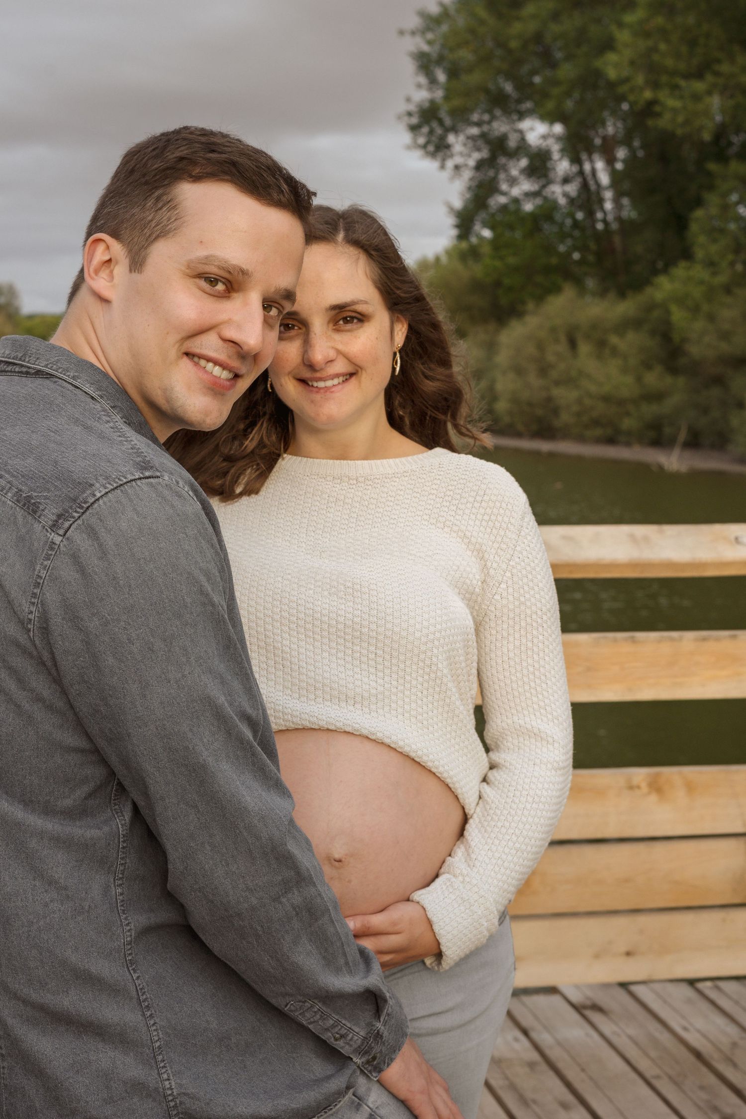 Happy couple posing outdoors on a wooden deck, pregnant woman cradling her baby bump beside her smiling partner.