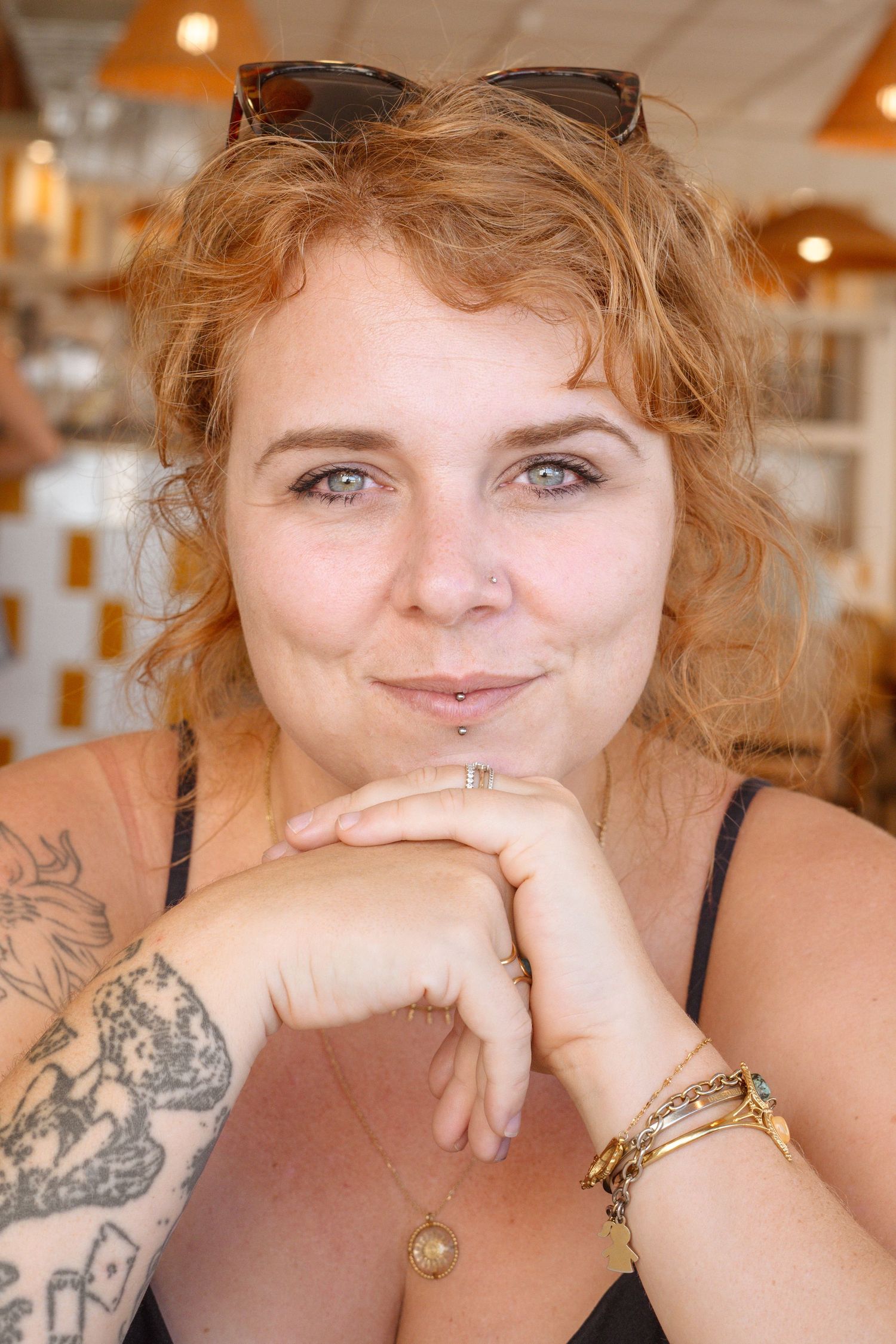 Smiling redhead woman with blue eyes, tattoos, and bracelets resting chin on hands in a bright cafe setting.