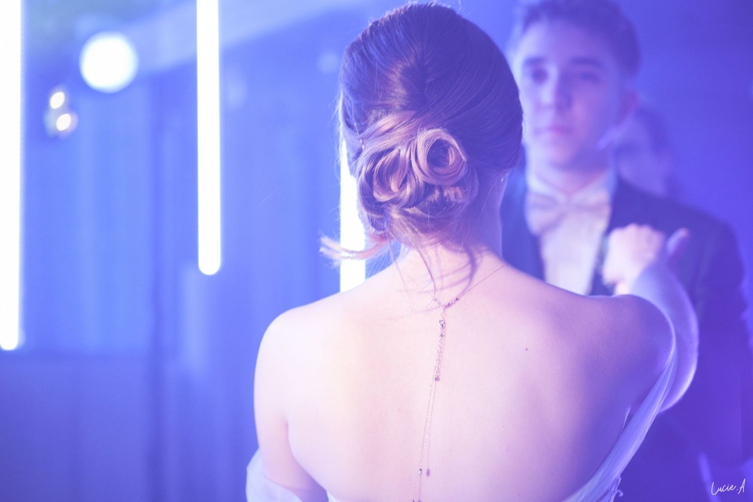 Une mariée, coiffée d'un chignon élégant et portant un délicat collier dans le dos, danse à la réception de mariage sous une lumière bleue.