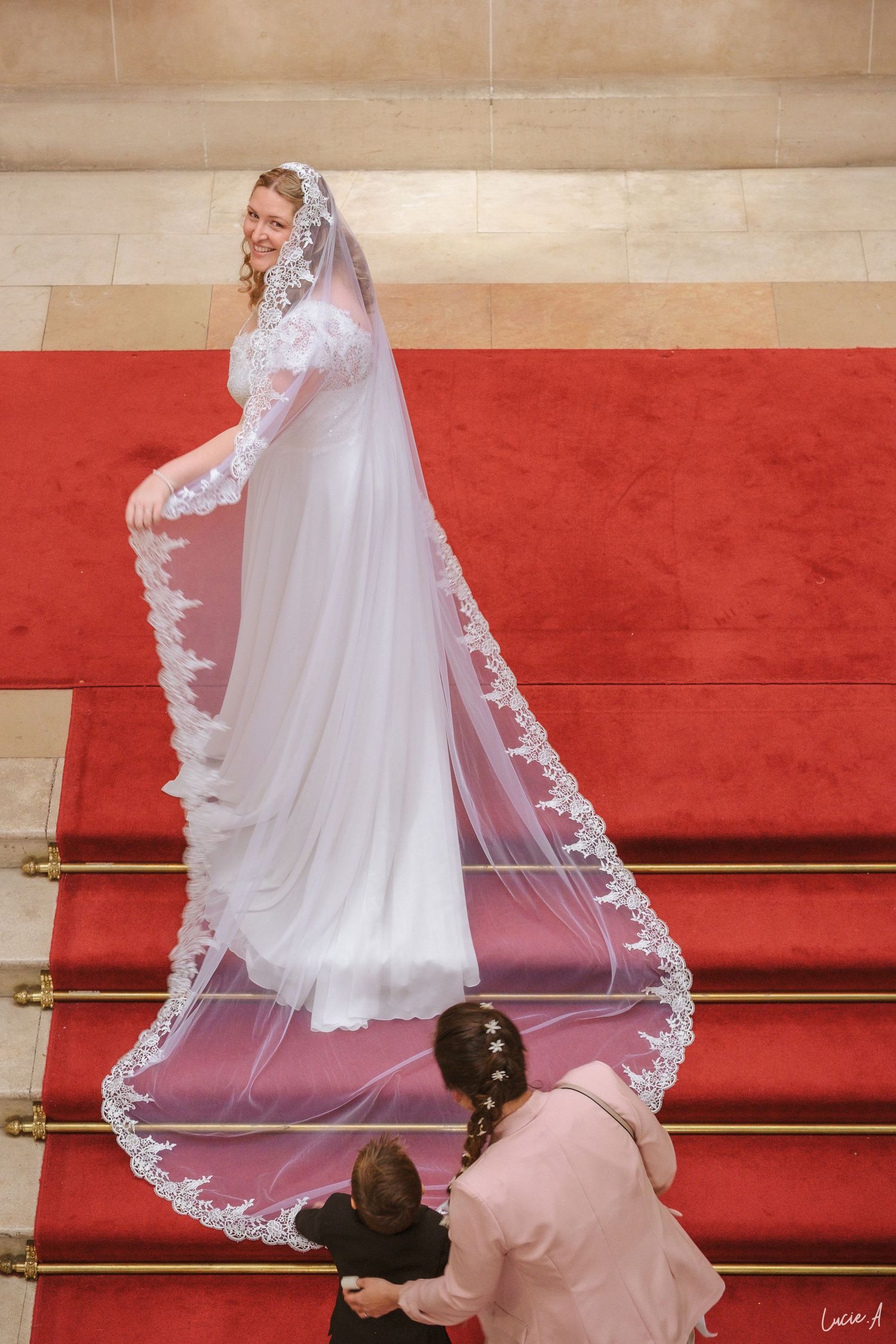 La mariée, vêtue d'une robe de dentelle blanche et d'un voile, descend un escalier recouvert de tapis rouge, assistée par sa témoin.