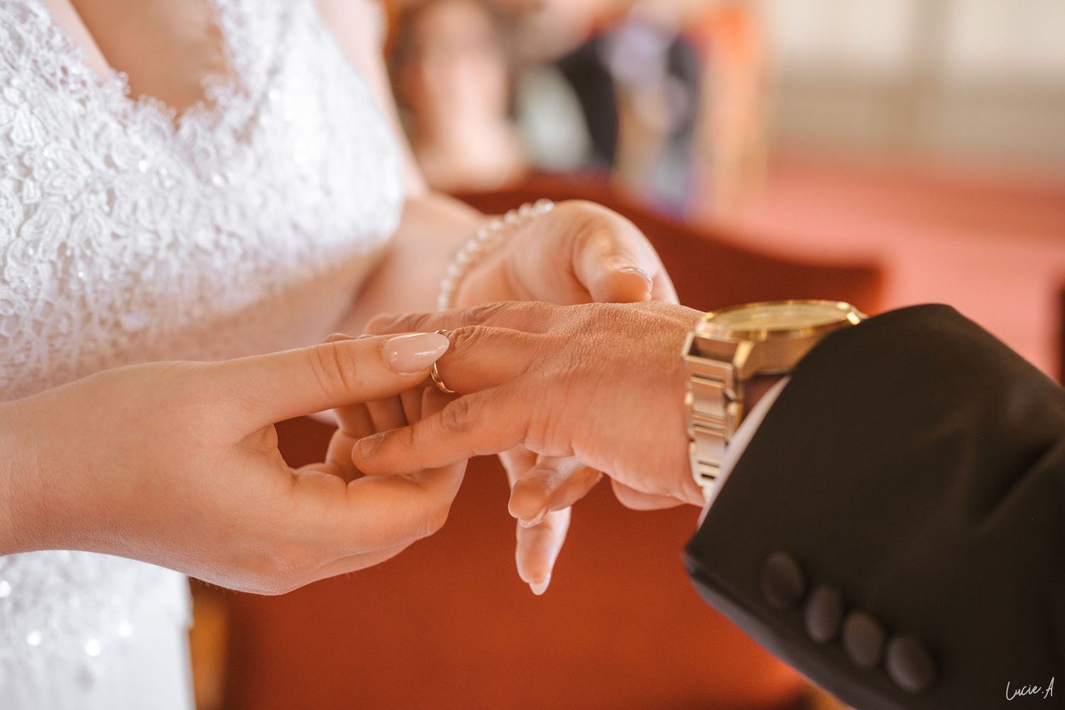 La mariée passe l'alliance au doigt du marié pendant la cérémonie ; le marié porte une montre en or et un costume sombre.