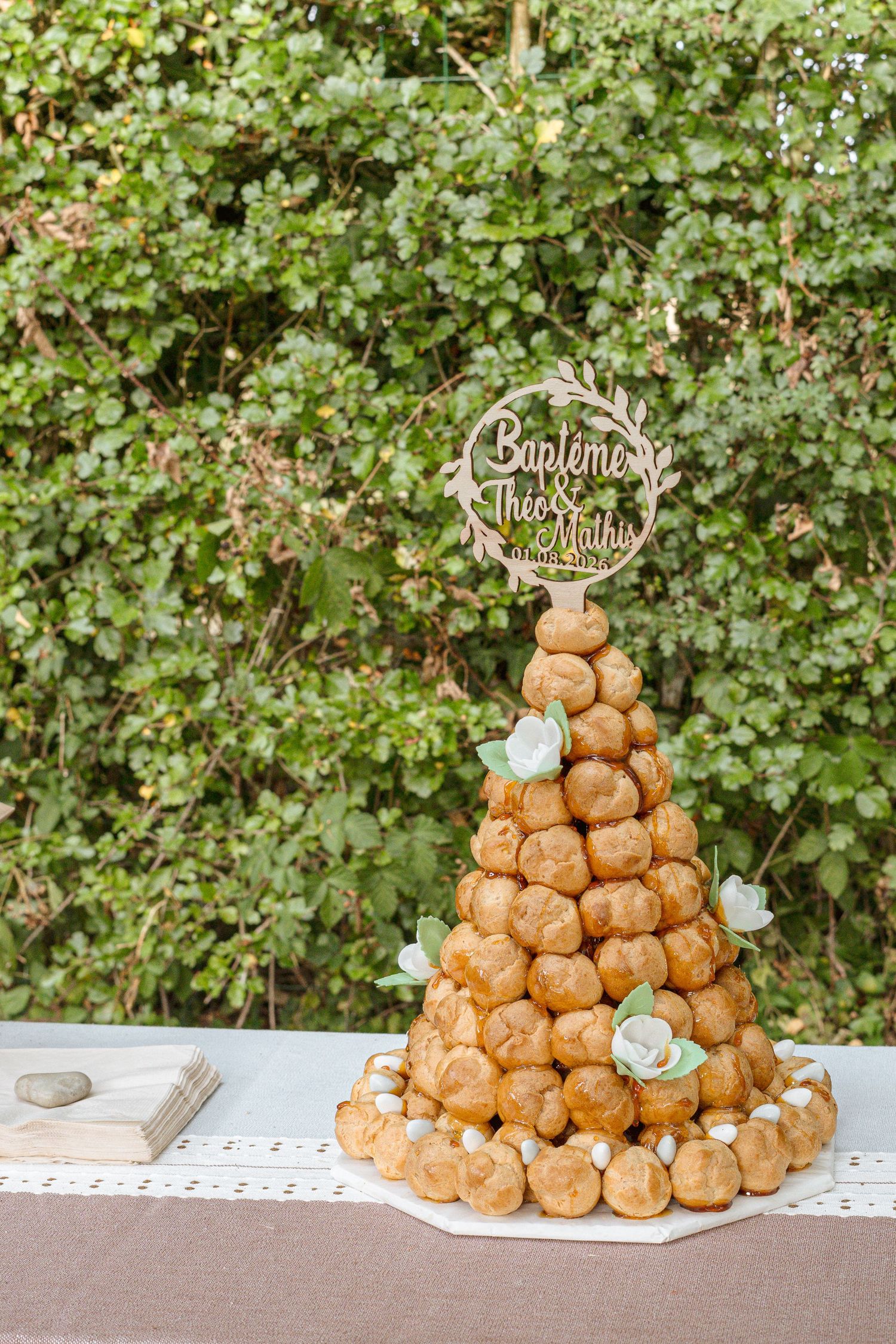 Elegant croquembouche tower decorated with cream puffs, white flowers, and a personalized wooden topper for a baptism celebration.