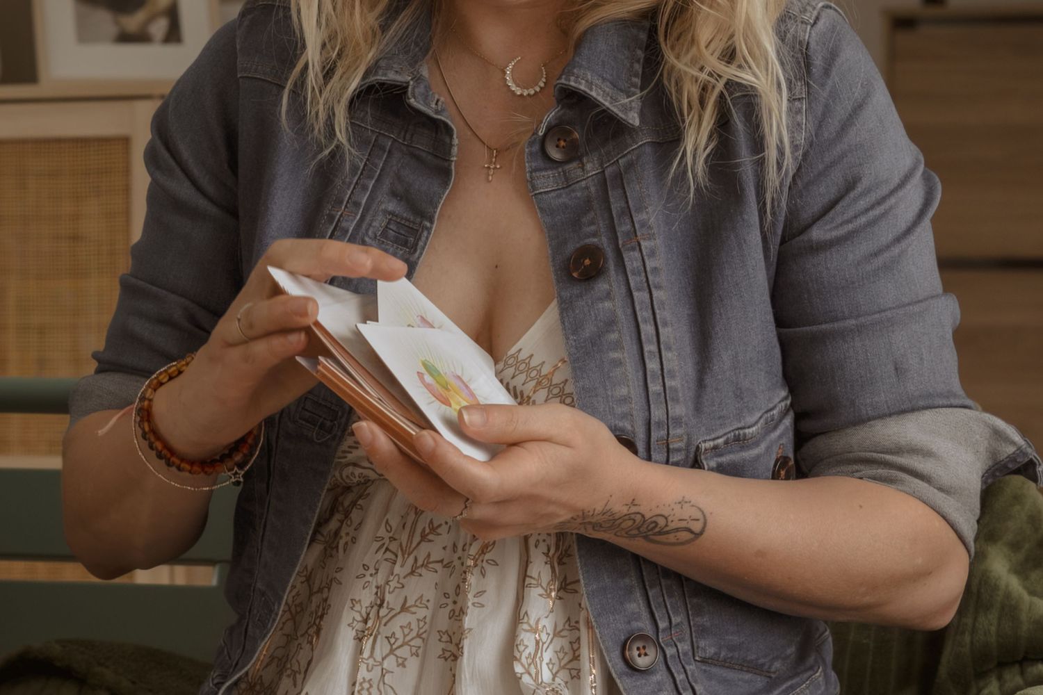 Woman in denim jacket with tattoo opening a small white gift box, wearing layered necklaces and bracelet.