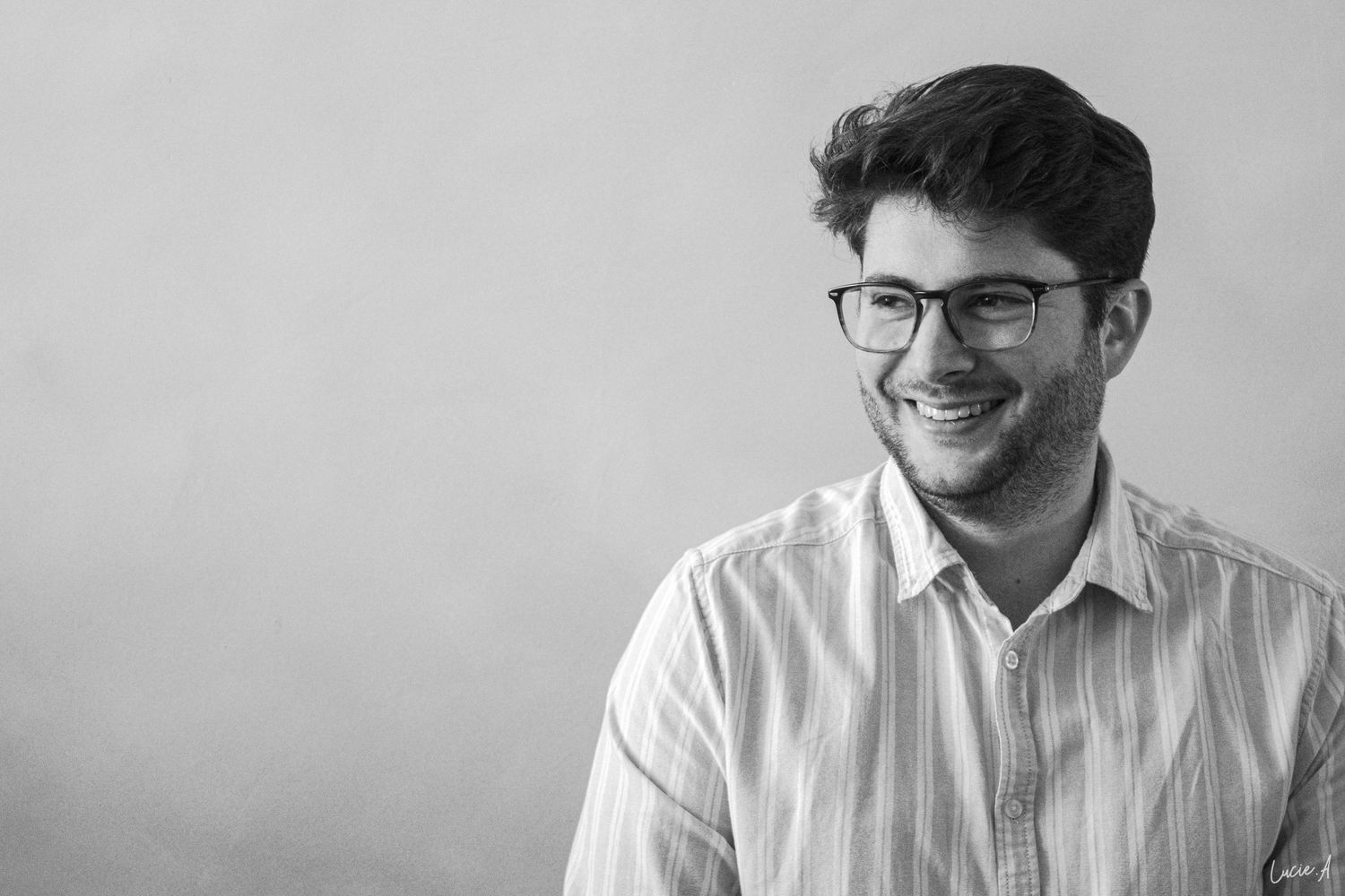 Jeune homme souriant avec des lunettes et une barbe portant une chemise rayée dans une photo de portrait en noir et blanc.