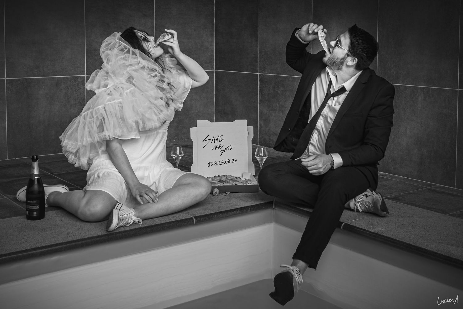 Photo en noir et blanc des mariés assis au bord d'une piscine, en train de boire et de manger une pizza