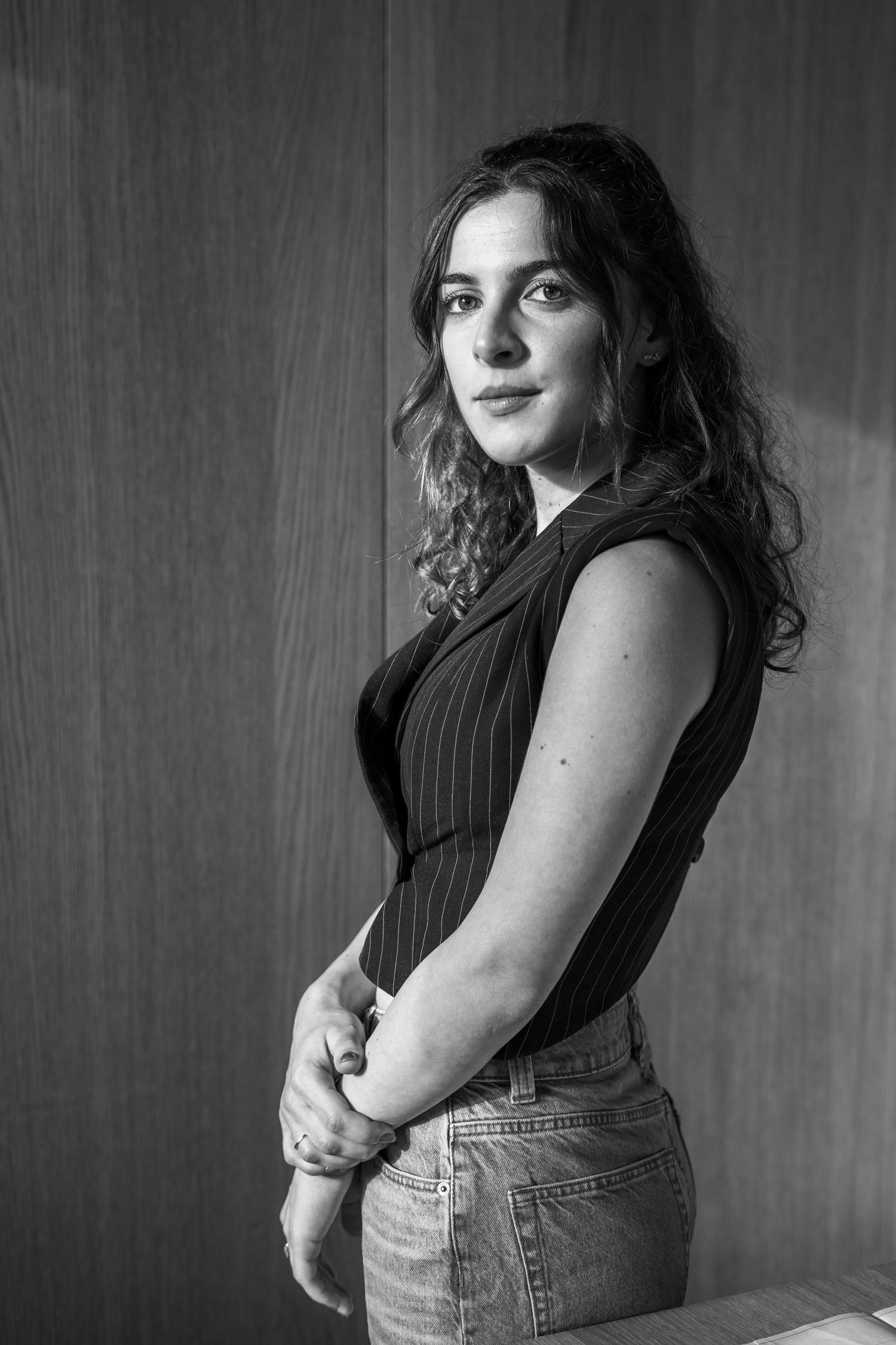 Black and white portrait of a young woman with wavy dark hair, wearing a pinstriped vest and jeans, posing against a wood-paneled wall.