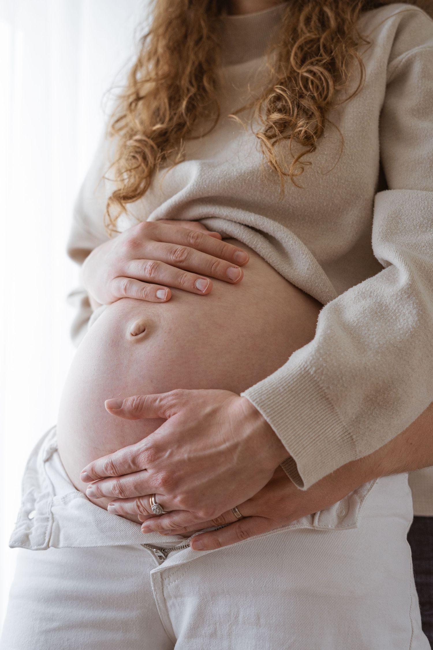 Pregnant woman with curly hair cradling her bare baby bump with both hands, wearing a beige sweater and white jeans.