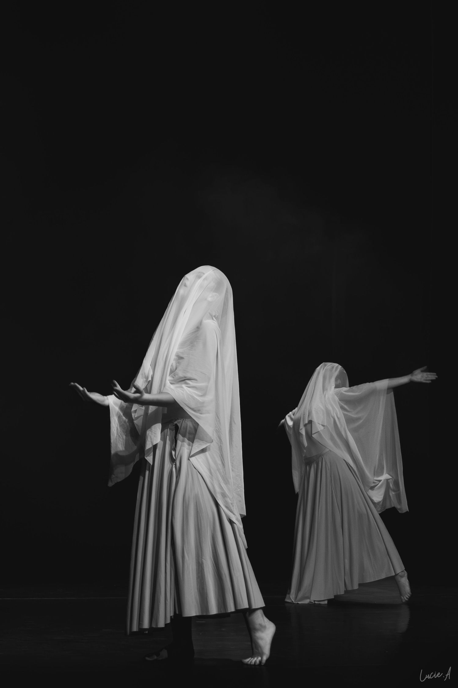 Deux silhouettes drapées de voiles blancs exécutent une danse expressive sur une scène sombre en noir et blanc.