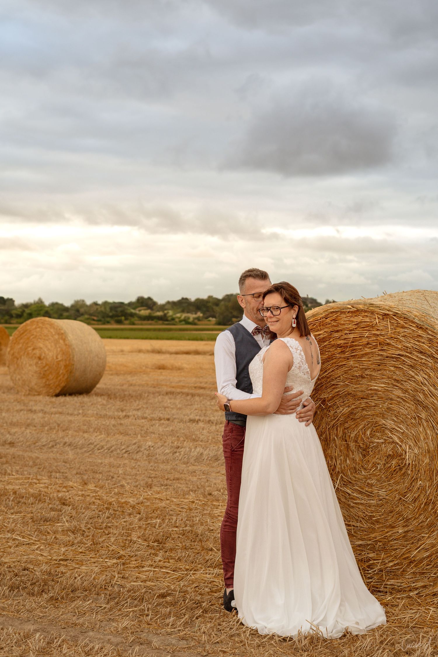 Un couple en tenue de mariage s'enlace près de bottes de foin dans un champ doré sous un ciel nuageux.