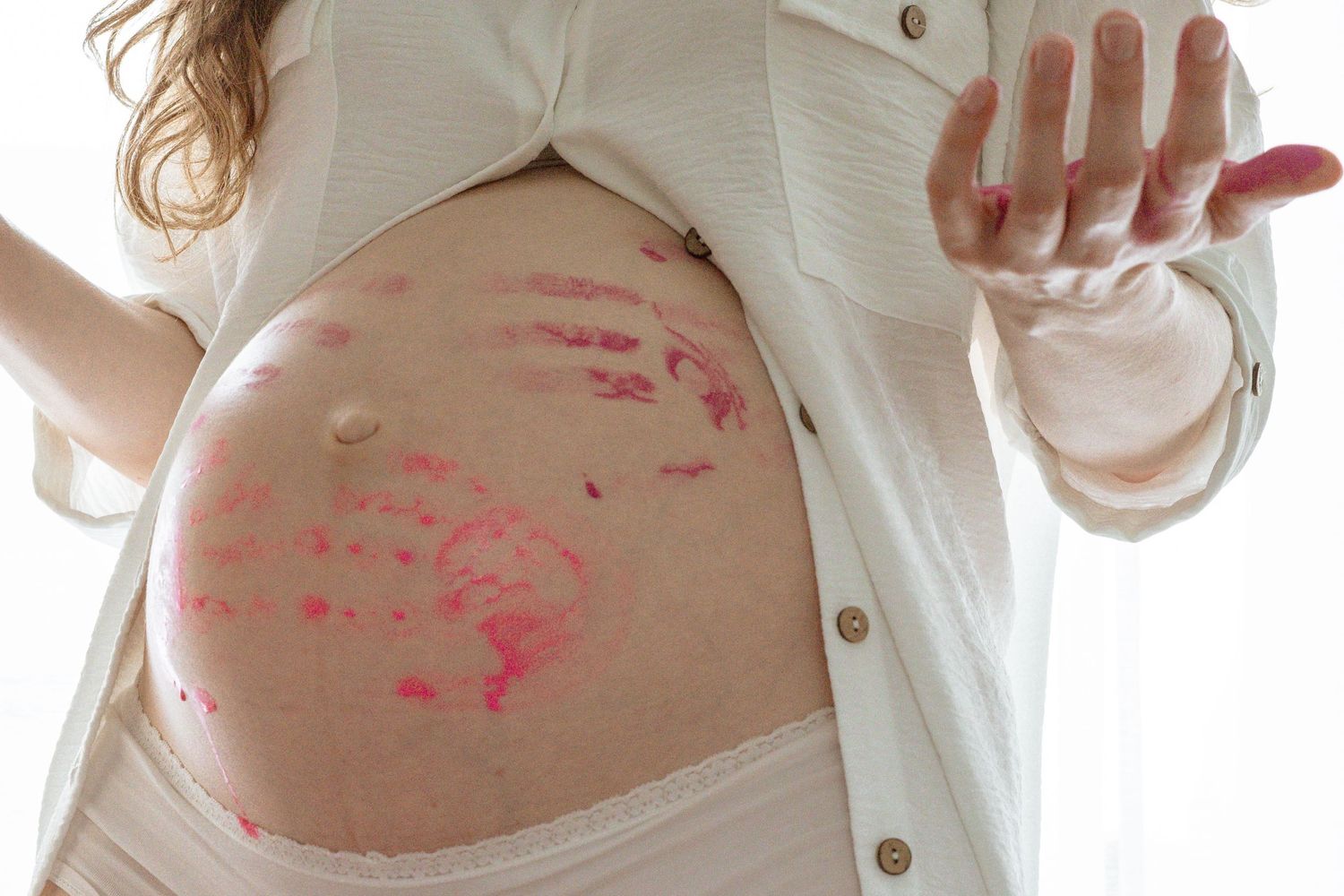 Pregnant woman with pink lipstick kiss marks on her bare baby bump, wearing an open white shirt.