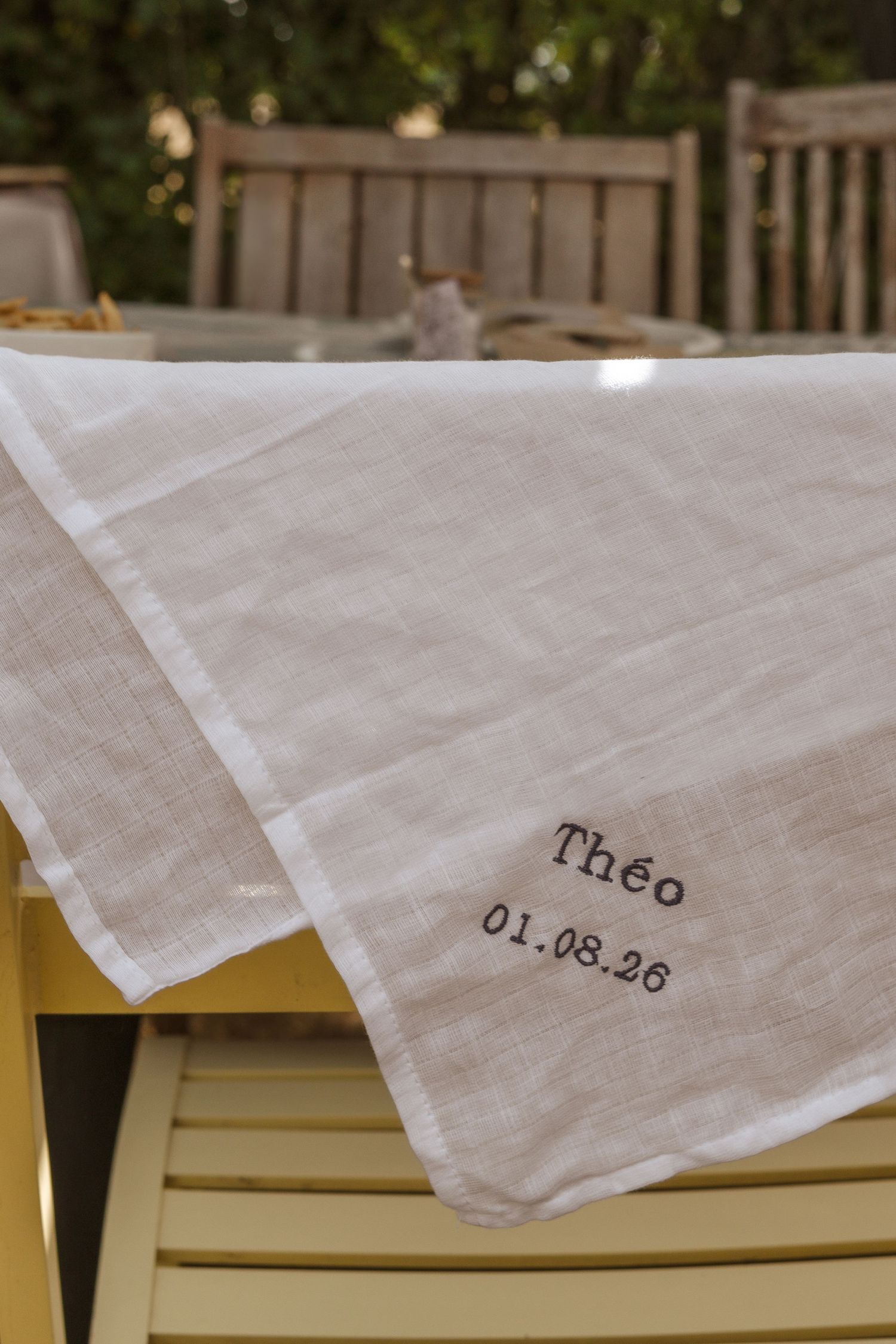 Serviette en lin blanc brodée du nom Théo et de la date 01.08.26, drapée sur une chaise jaune à l'extérieur.