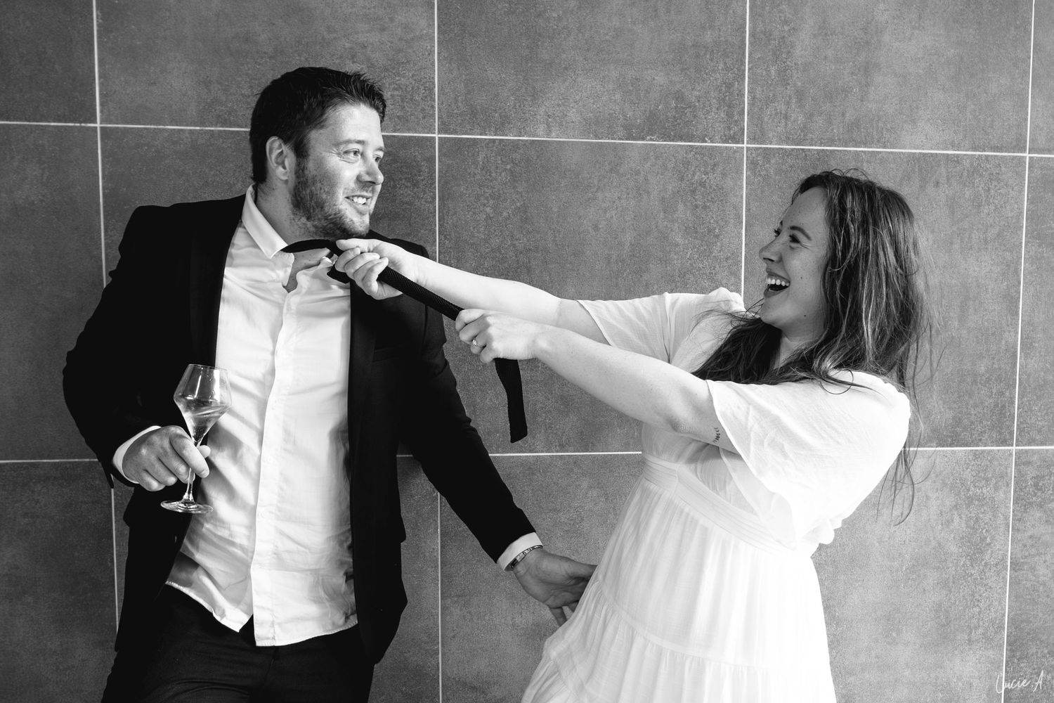 Photo en noir et blanc d'un couple riant, la femme saisissant en plaisantant la cravate de l'homme tandis qu'il tient un verre de vin.