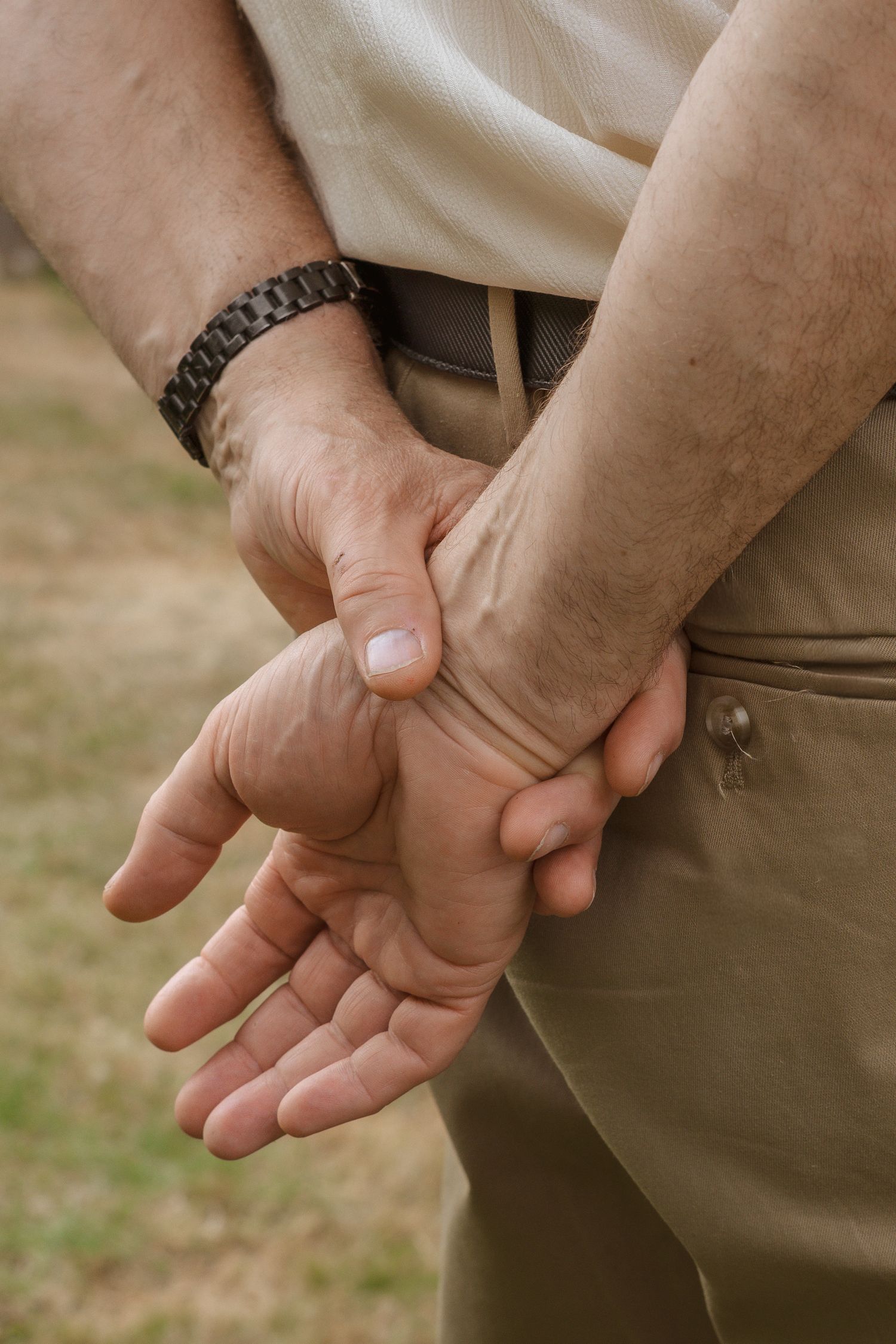 Close-up of a man holding his wrist behind his back, wearing khaki pants and a metal bracelet outdoors.
