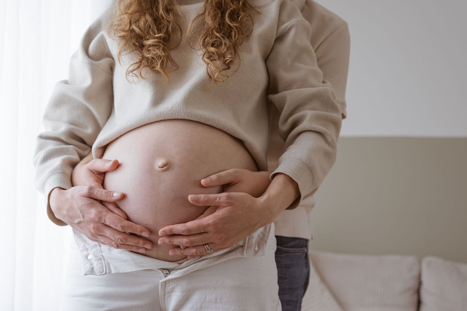 Pregnant woman in beige sweater cradling her bare baby bump with both hands in a bright room.