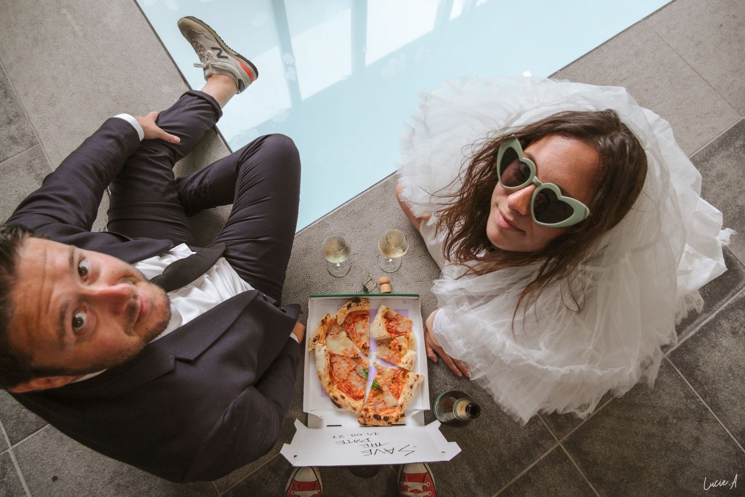 La mariée, vêtue d'une robe blanche et de lunettes de soleil vertes, est assise par terre avec le marié, partageant une boîte à pizza.