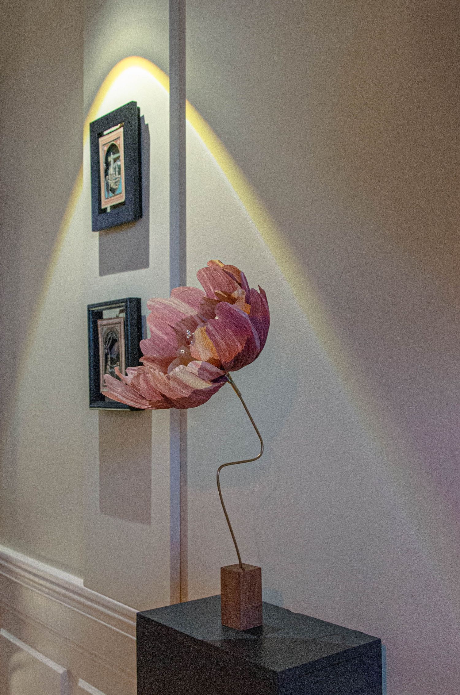 Sculpture de fleur de lotus rose sur un socle en bois, exposée sur un piédestal sombre à côté d'une œuvre d'art encadrée.