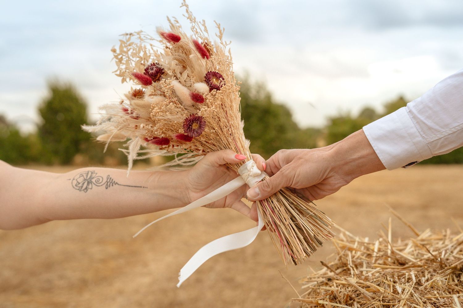 Deux mains échangent un bouquet rustique de fleurs séchées, aux fleurs rouges nouées d'un ruban blanc, près d'une botte de foin.