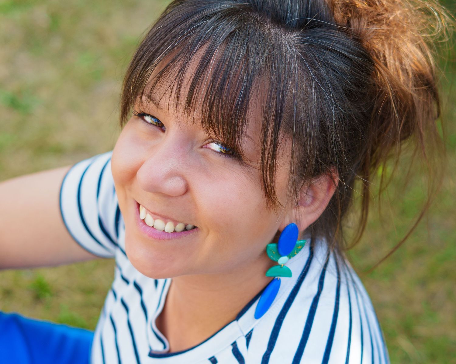 Smiling young woman with bangs and updo wearing blue and green earrings and striped shirt outdoors.