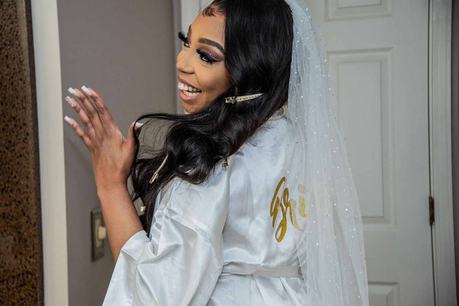 Radiant bride in white satin robe and veil smiling while getting ready on her wedding day.
