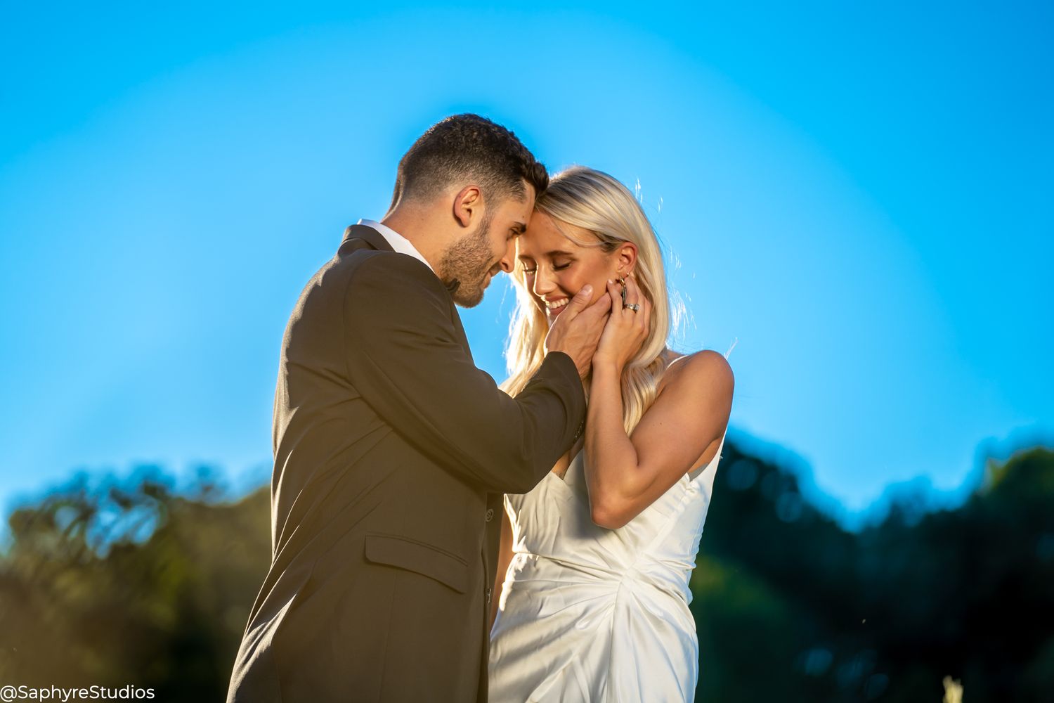 Groom gently cupping brides face in an intimate moment against a vivid blue sky.
