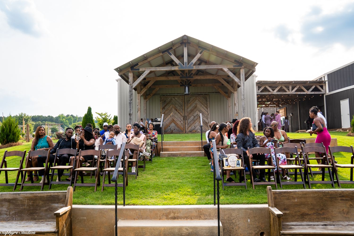 Outdoor rustic barn wedding ceremony with guests seated in rows on a lush green lawn.