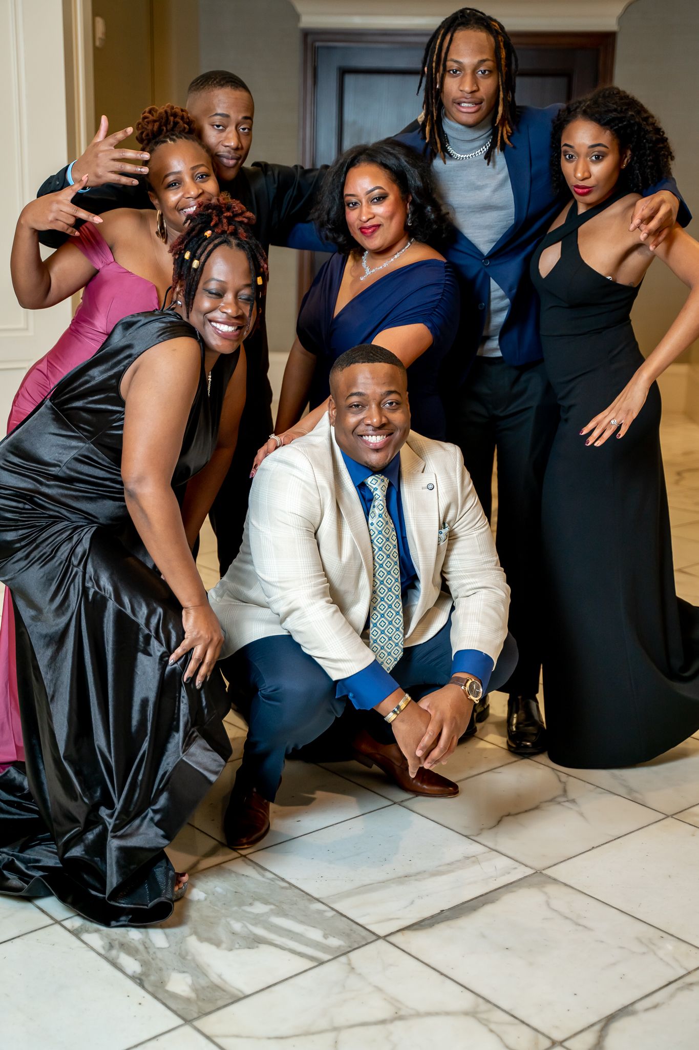 Group of seven elegantly dressed friends posing together at a formal event in a marble-floored venue.