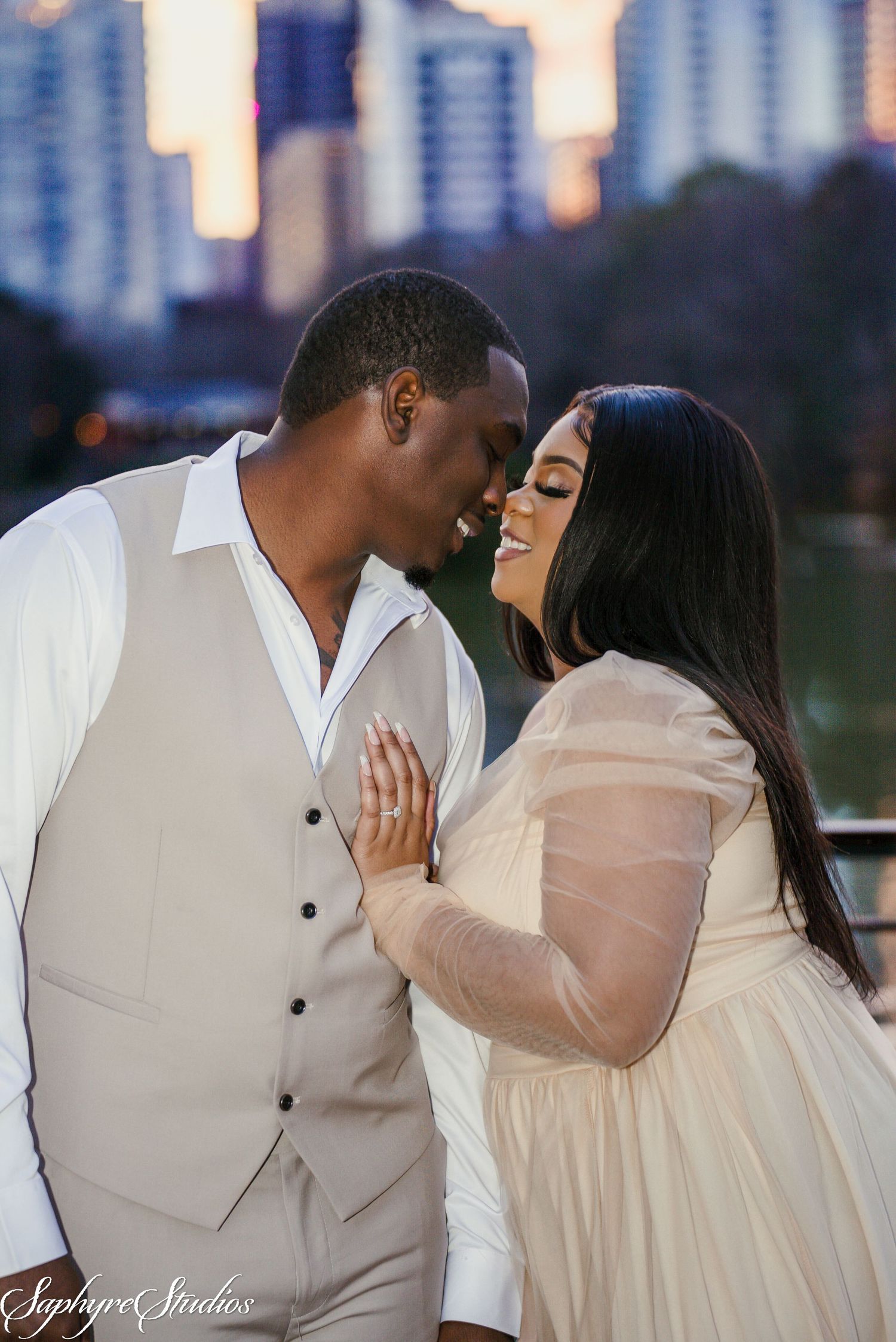 Engaged couple sharing a tender moment in white attire with city skyline backdrop at golden hour.