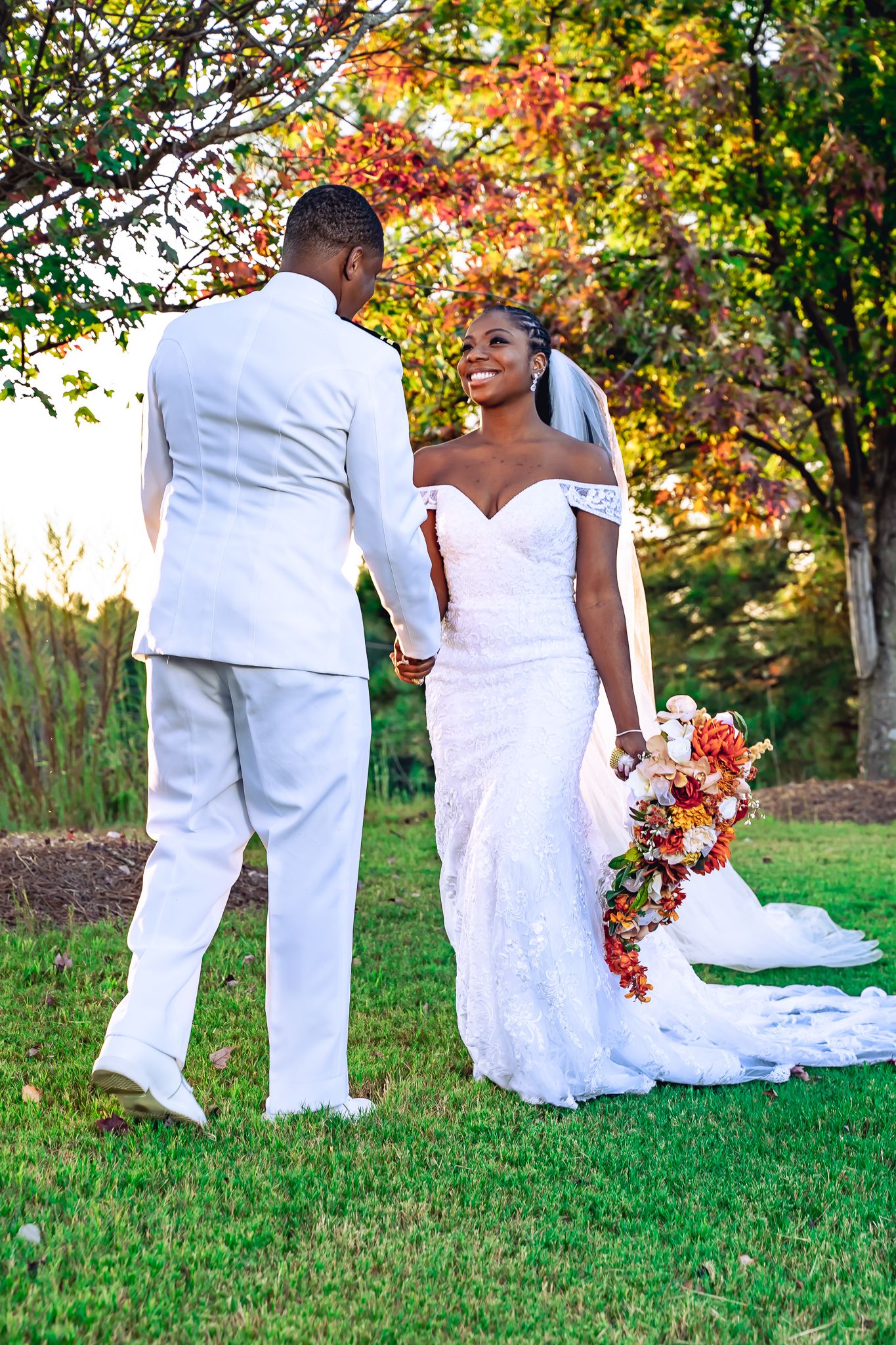 Bride in white lace gown holding cascading bouquet walking hand-in-hand with groom among autumn foliage.