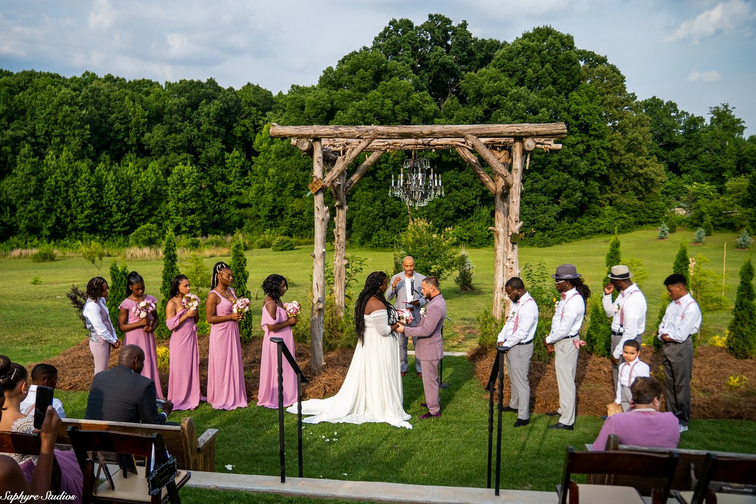 Wedding party stands at rustic wooden arch altar during outdoor ceremony as guests watch from wooden chairs.