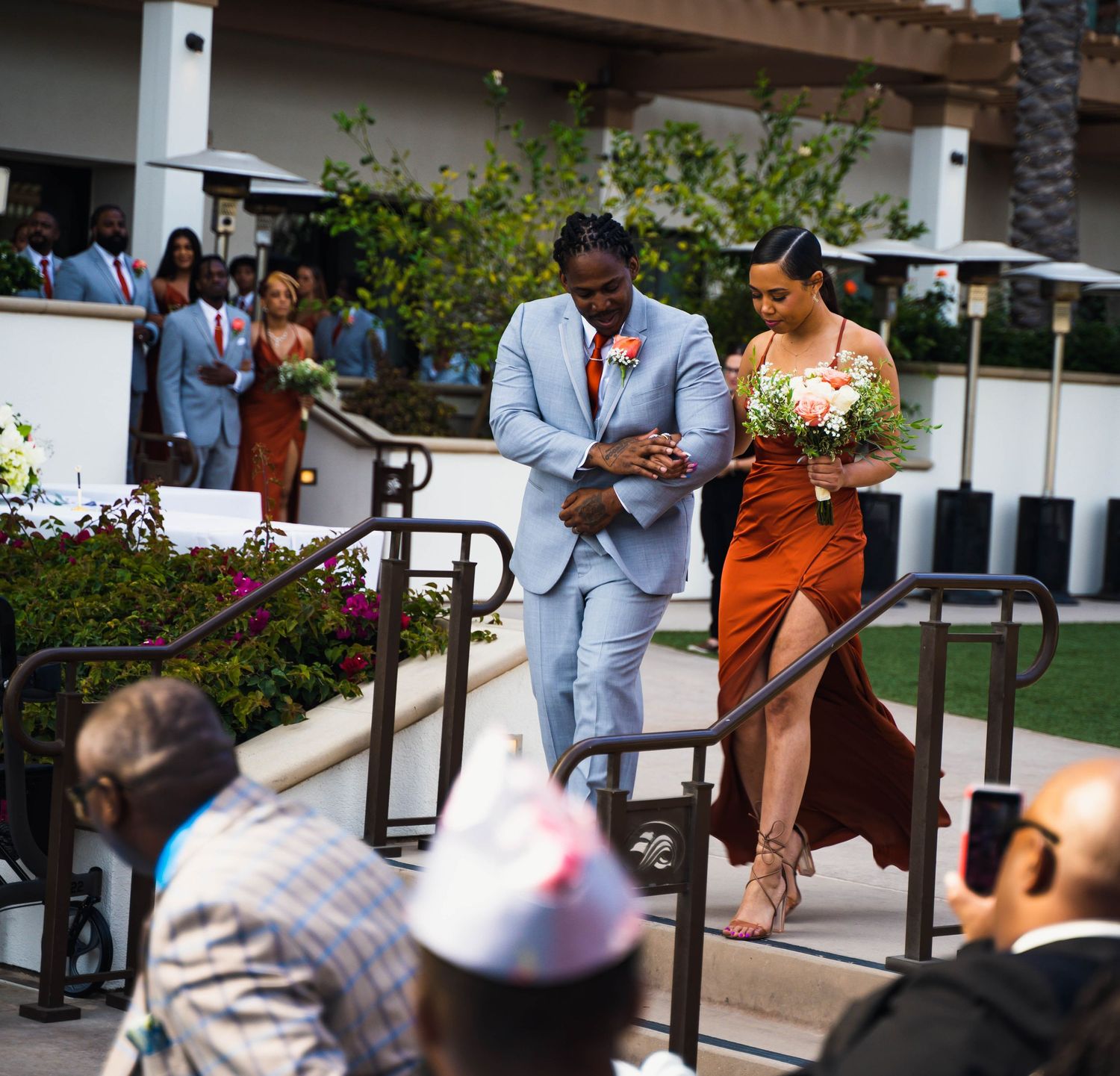 Groomsman in light blue suit walks bridesmaid in burnt orange dress down wedding ceremony steps.