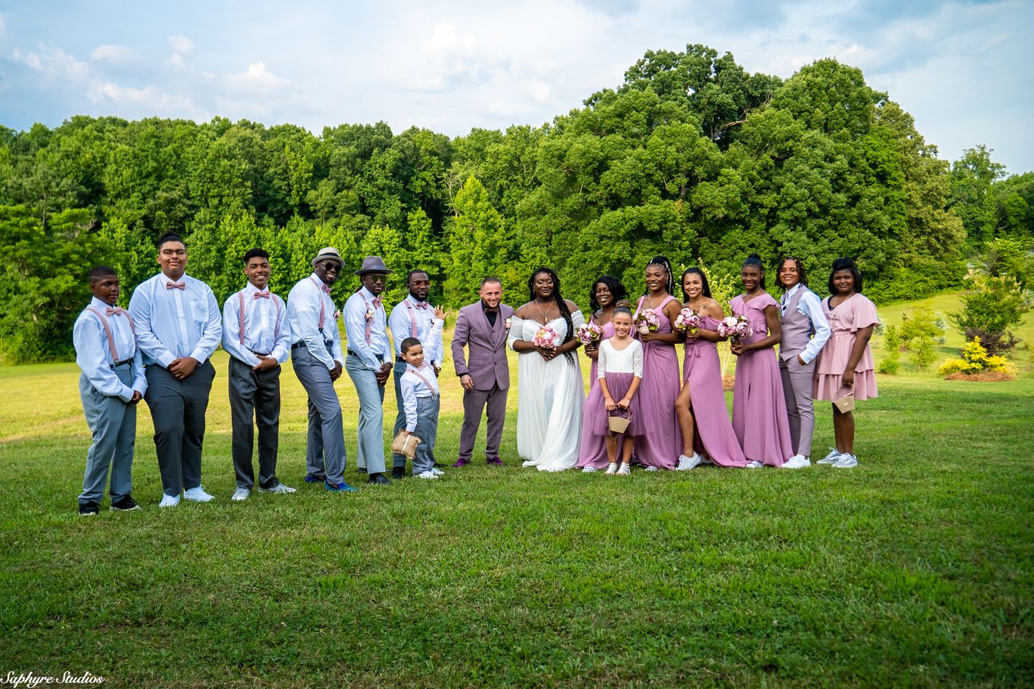 Large wedding party poses outdoors on green lawn with lush trees, groomsmen in gray suits and bridesmaids in mauve dresses.
