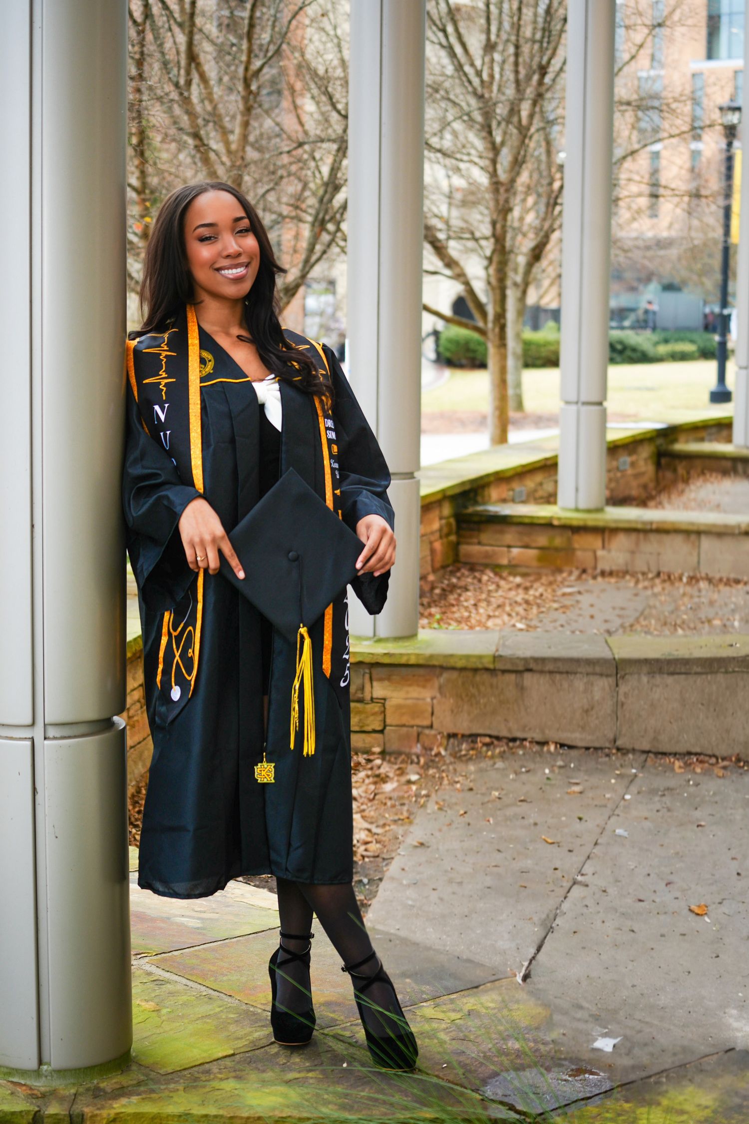 Smiling graduate in black and gold regalia leaning against column on campus.