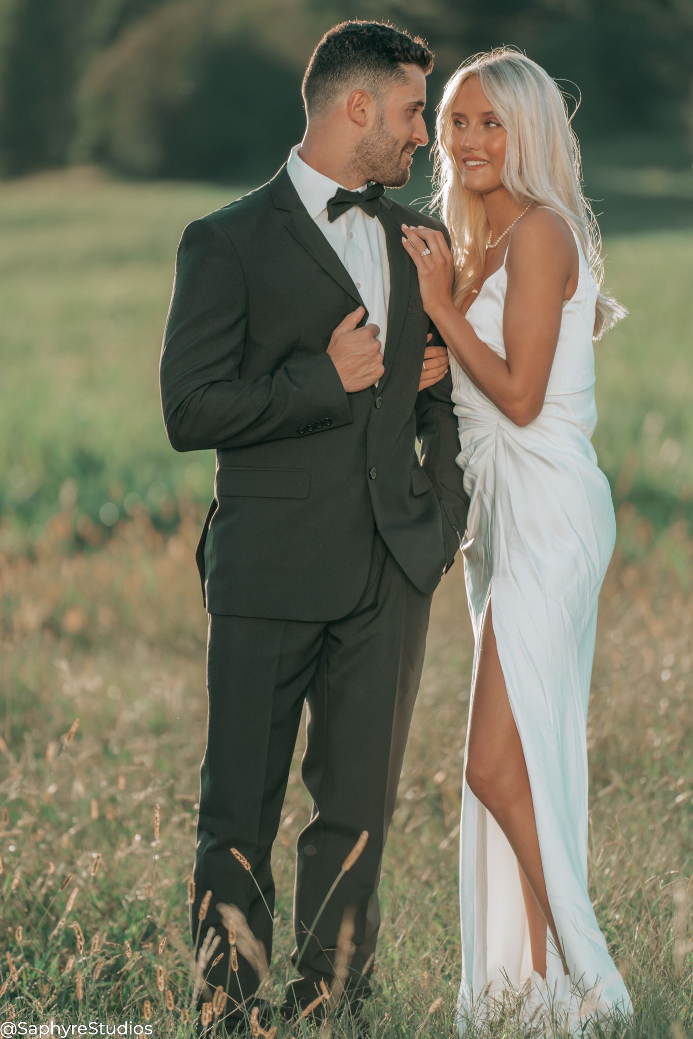 Stunning blonde bride in white slit gown smiling with groom in black tuxedo in golden field.