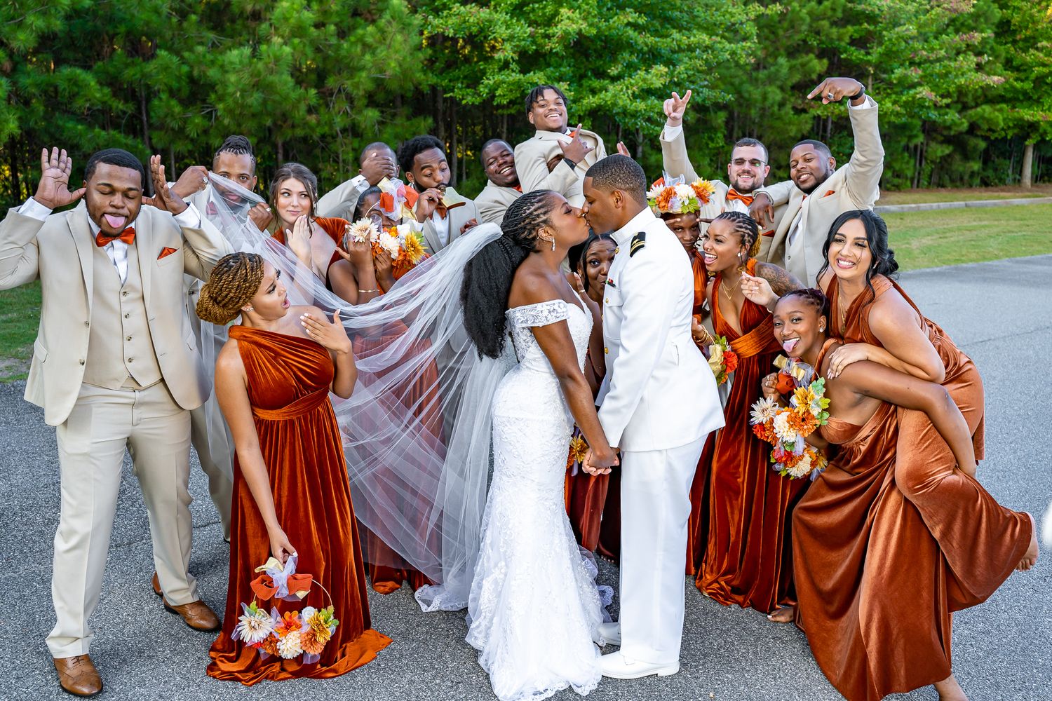 Wedding party celebrating as bride and groom kiss, bridesmaids in rust velvet and groomsmen in beige suits.