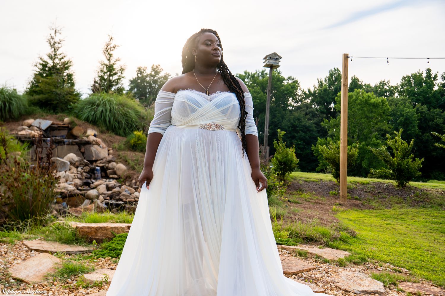 Bride in flowing white off-shoulder wedding gown stands in garden with waterfall and string lights in background.
