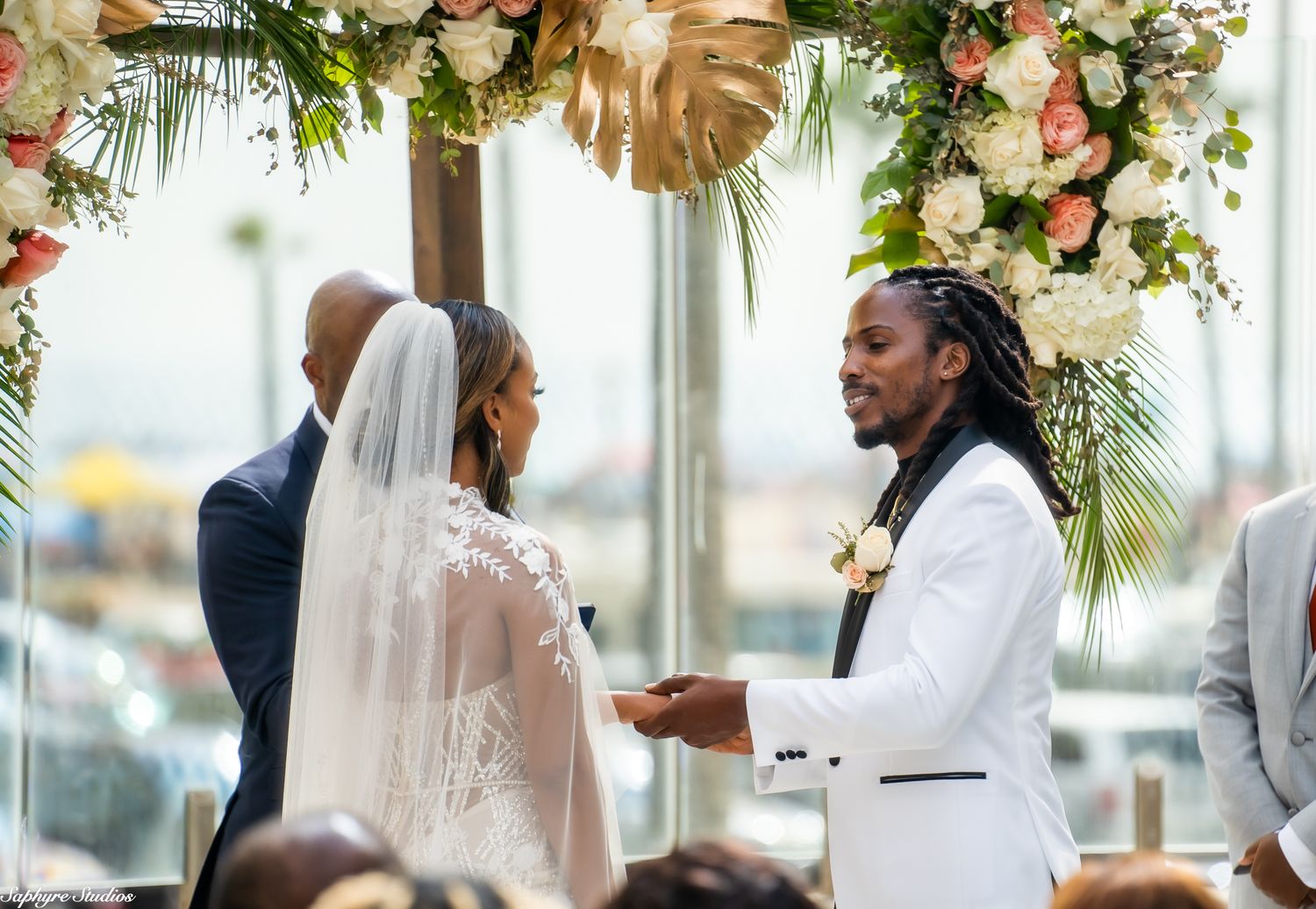Smiling groom in white suit holds brides hands under floral arch at outdoor wedding ceremony.