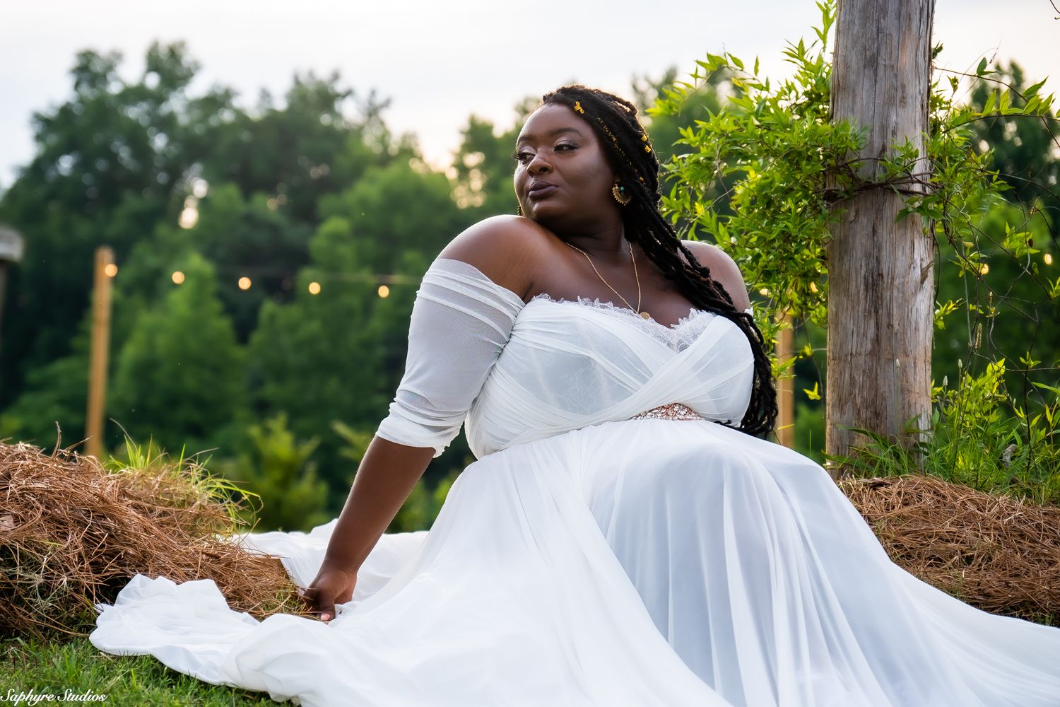 Bride in white flowing wedding gown reclines gracefully on hay bale in vineyard with string lights at dusk.
