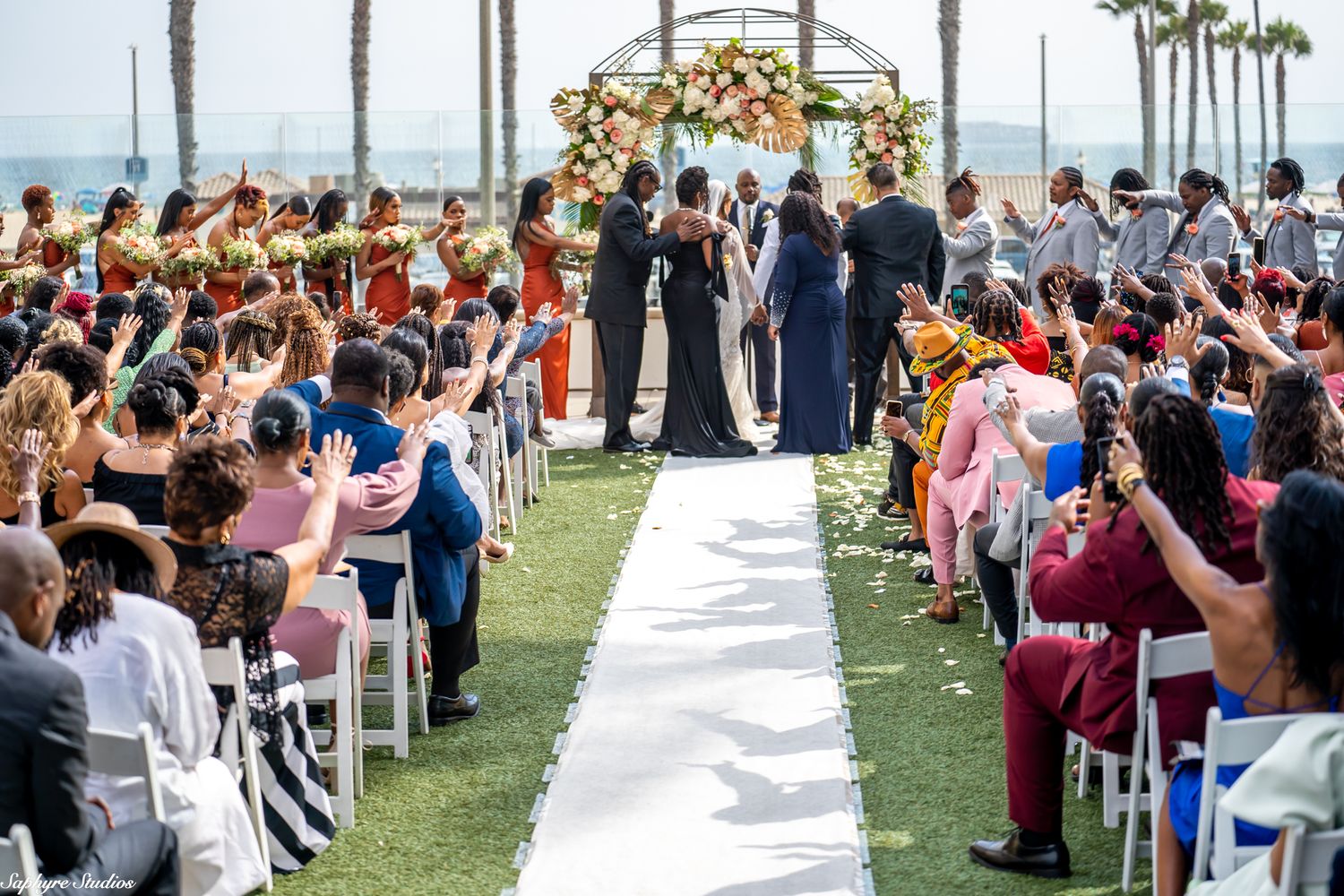 Wedding guests raise hands in celebration as couple stands at altar during outdoor ceremony.