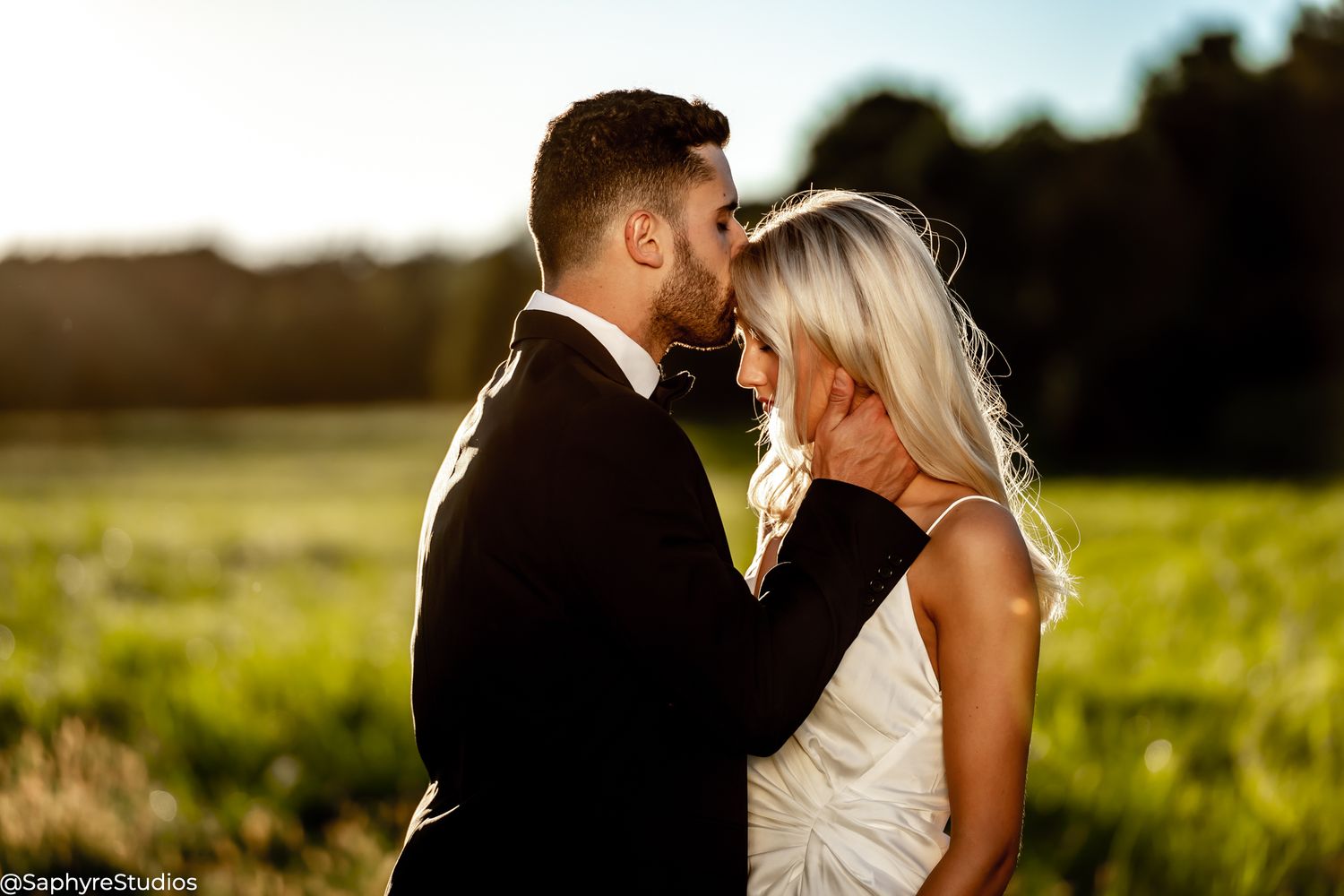 Groom leaning in to kiss bride in a golden sunlit green field at sunset.