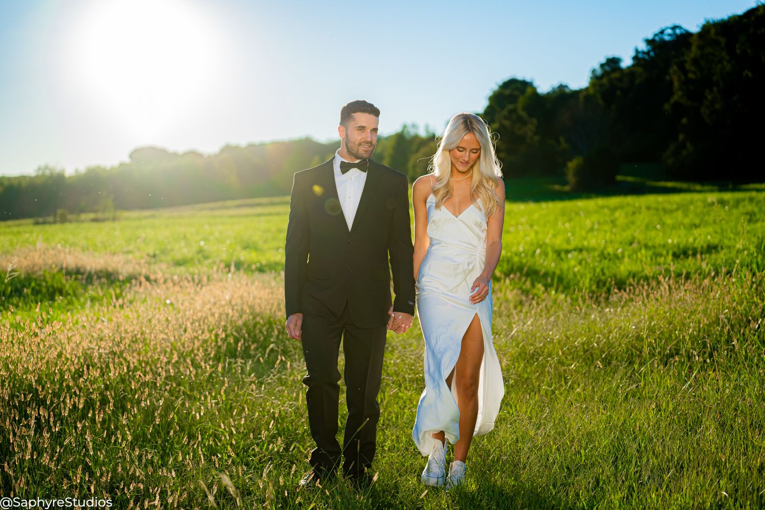 Wedding couple holding hands strolling across lush green meadow in bright sunlight.