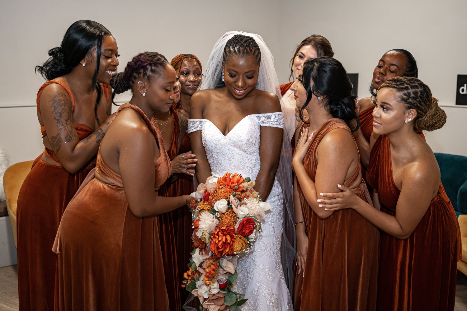 Bride in white lace gown surrounded by bridesmaids in rust orange dresses admiring her bouquet of fall flowers.