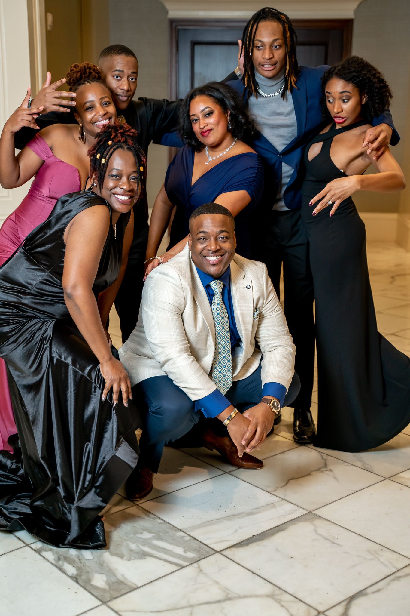 Seven friends in formal attire striking fun poses together at an upscale indoor celebration.