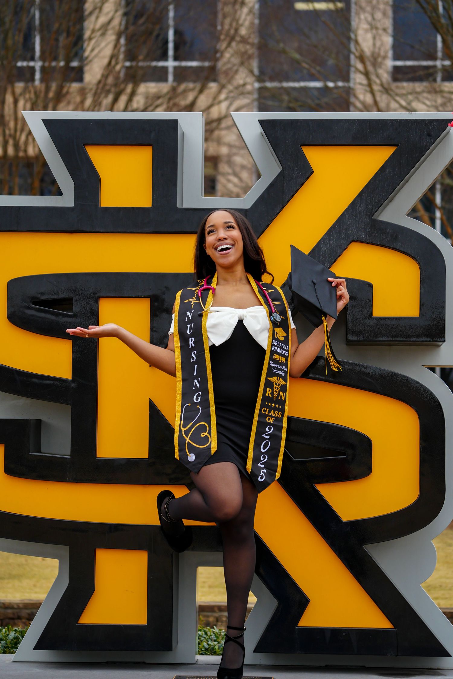 Joyful nursing graduate poses with cap and sash beside KSU large campus sign at Kennesaw State University.