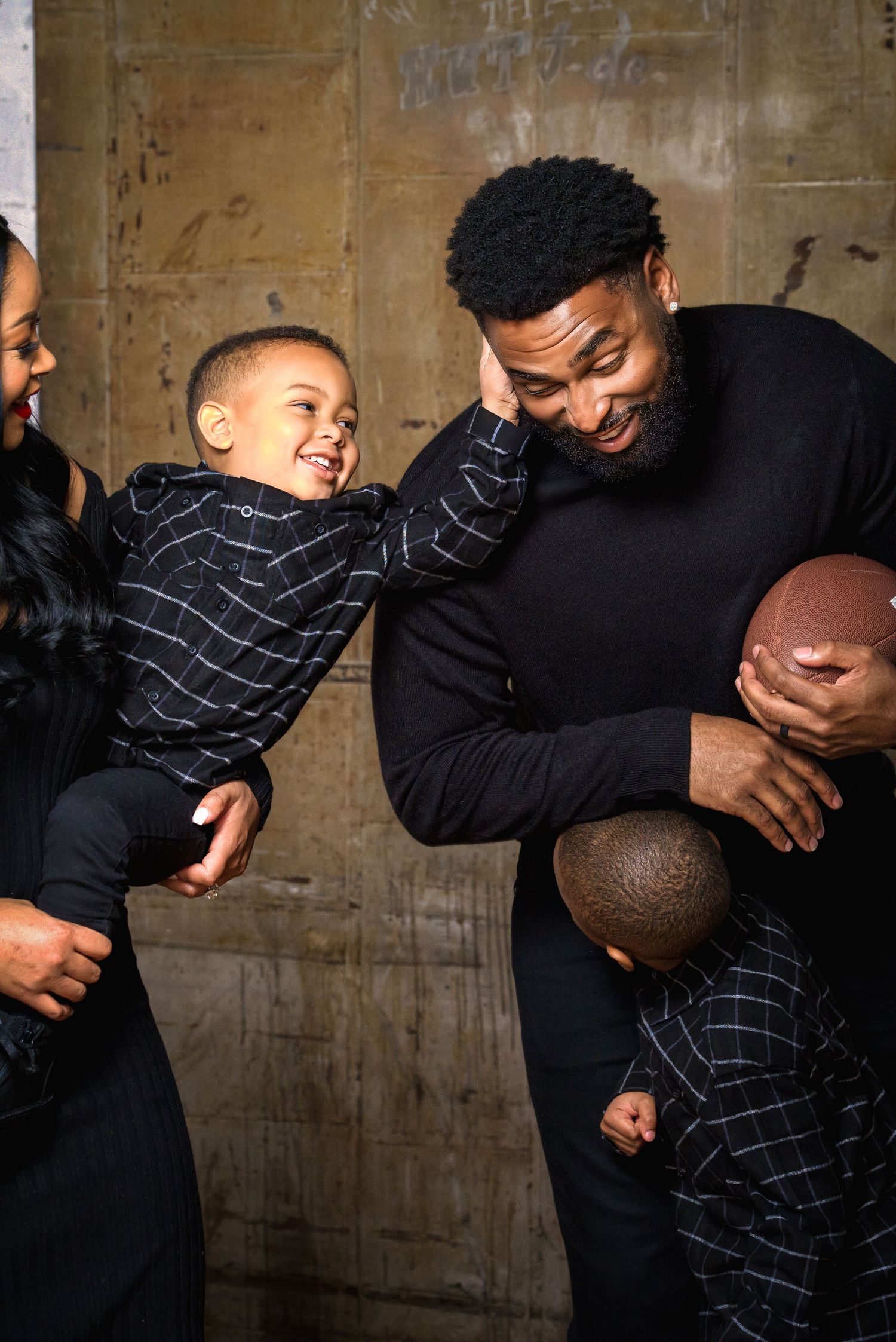 Joyful Black family in matching black outfits, father holding football and laughing toddler son.