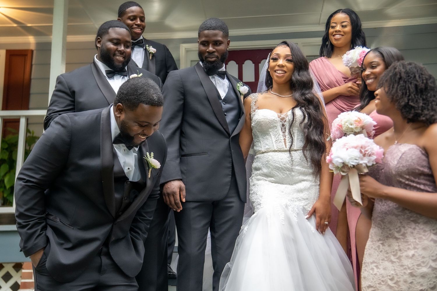 Groom and groomsmen in dark suits laugh with bride and bridesmaids during wedding party photos.