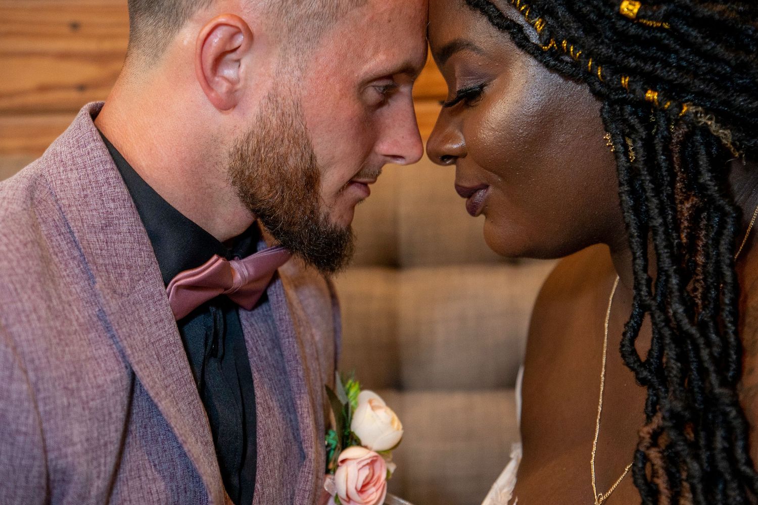 Interracial bride and groom touch foreheads intimately, groom in mauve suit with rose boutonniere at wedding.