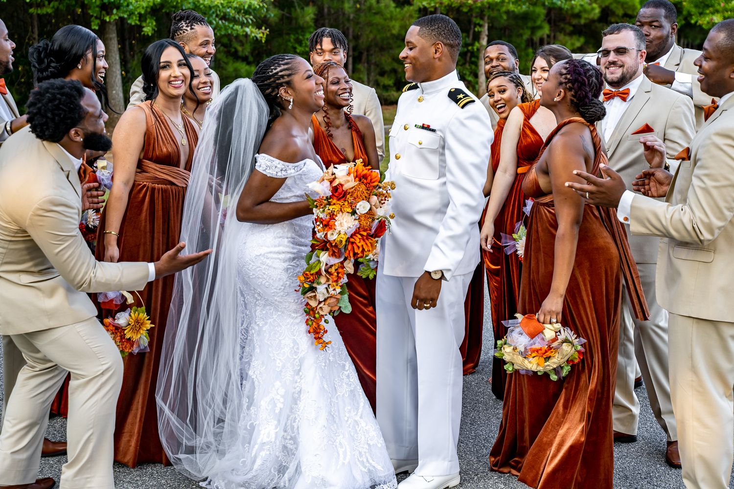 Joyful wedding party surrounding bride in white lace gown and military groom in white uniform outdoors.