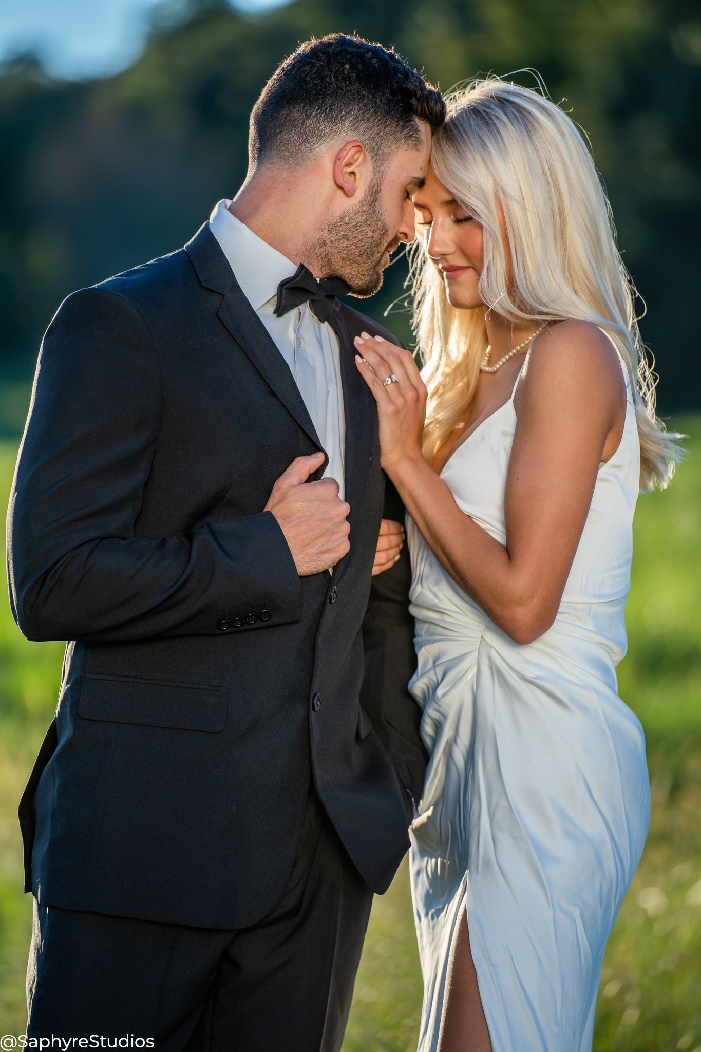 Romantic bride and groom in black tuxedo sharing intimate moment in sun-kissed outdoor field.