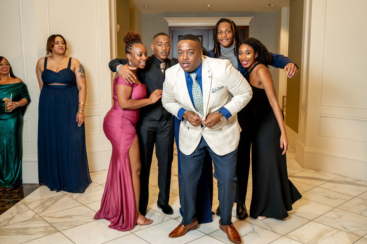 Formally dressed group posing playfully in a hallway, man in white blazer stands in front.